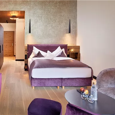 A modern hotel room with a large bed and a cozy seating area. The decor is in purple and wood, with soft lighting.