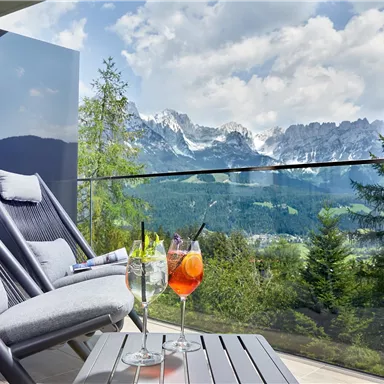 A balcony with two lounge chairs and a view of the mountains. On the table are two cocktails and the surroundings are green and picturesque.