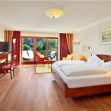 A cozy hotel room with two beds and wooden furniture. Large windows offer a view of a balcony with seating.