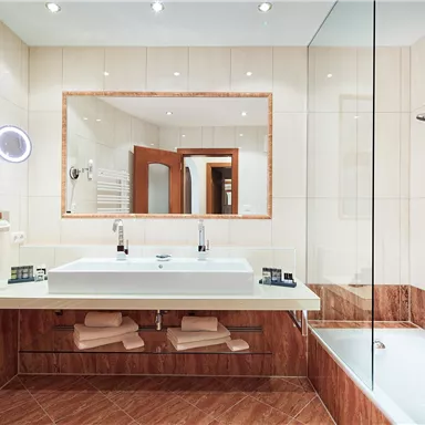 A modern bathroom with a large double sink, a bathtub, and a separate shower cabin. The walls are tiled in light colors and the lighting is pleasant.
