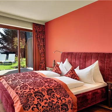 A modern bedroom with a red bed and sleek bedding. Large windows provide a view of the garden and the outdoor area.