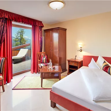 A cozy bedroom with a red bed and wooden furniture. Through the window, there is a beautiful view of the terrace and the surrounding nature.