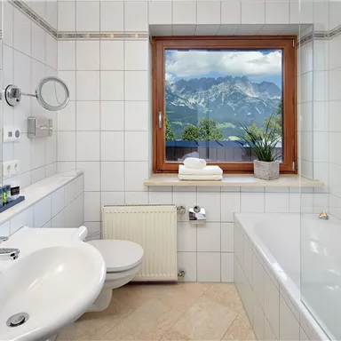 A modern bathroom with white tiles and natural light. Through the window, one can see an impressive mountain landscape.