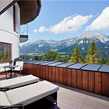 A modern terrace with loungers and a table, surrounded by majestic mountains. The sky is clear and blue, and the view is breathtaking.