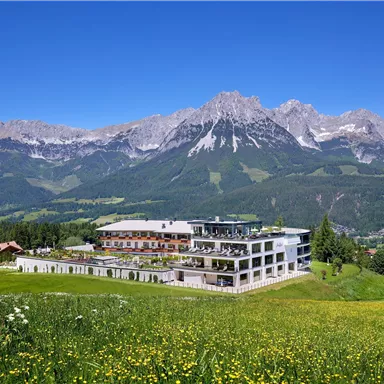 A modern building complex surrounded by green meadows and mountains. The clear blue sky gives the scene a fresh atmosphere.