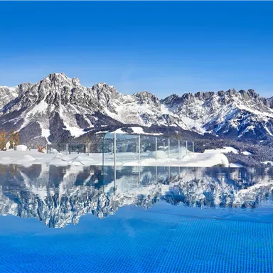 Ein atemberaubender Blick auf schneebedeckte Berge mit einem glitzernden Schwimmbad im Vordergrund. Der klare blaue Himmel spiegelt sich im Wasser wider.