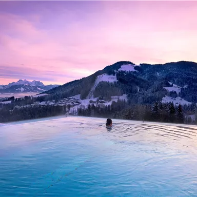 An infinity pool overlooking snow-capped mountains at twilight. The landscape is characterized by soft colors and a calm sky.