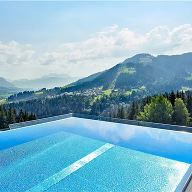 An infinity pool with clear water overlooks a beautiful mountain landscape. The surroundings are green and wooded, with gentle hills and a blue sky.