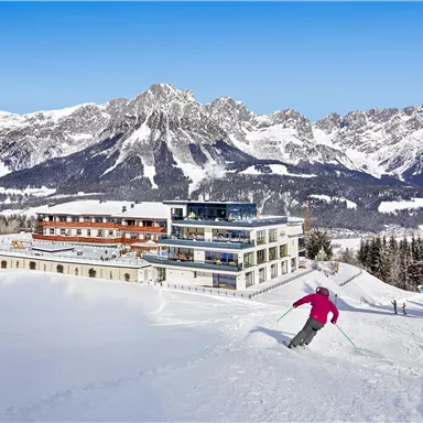 Eine verschneite Berglandschaft mit einem modernen Hotel und majestätischen Bergen im Hintergrund. Im Vordergrund fährt ein Skifahrer die Piste hinunter.