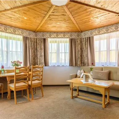 Ein gemütlicher Wohnraum mit einem runden Tisch und Stühlen. An den Fenstern hängen Vorhänge, und eine Couch lädt zum Verweilen ein.