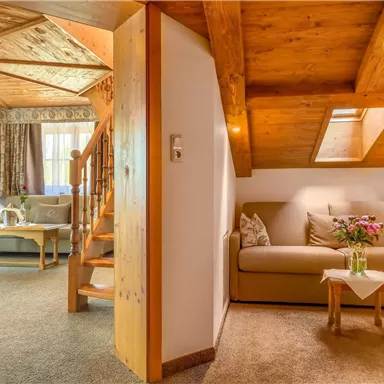 Eine gemütliche Wohnstube mit einem Sofa und einem Tisch. Die Holzbalkendecke und die warmen Farben schaffen eine einladende Atmosphäre.