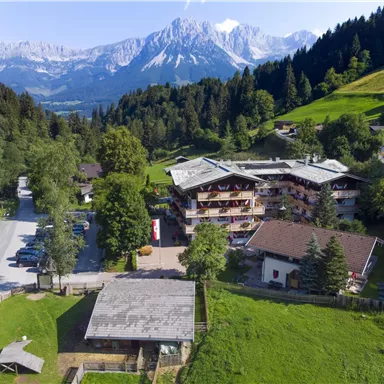 Ein malerisches Berghotel umgeben von grünen Wiesen und dichten Wäldern. Im Hintergrund erheben sich majestätische Berge unter einem klaren Himmel.