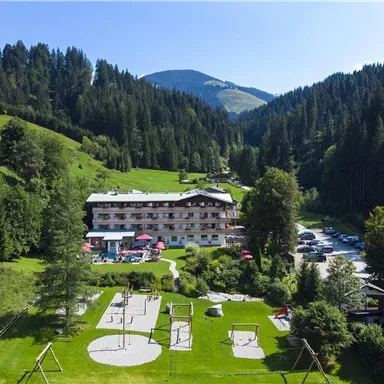 Ein malerisches Hotel in einer grünen Berglandschaft. Im Vordergrund befinden sich Spielmöglichkeiten für Kinder und eine schöne Rasenfläche.