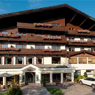 Ein charmantes Hotelgebäude mit Balkonen und Blumenschmuck. Die sonnige Umgebung und die Berglandschaft verleihen dem Ort eine einladende Atmosphäre.