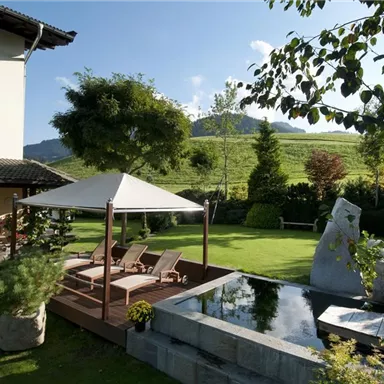 Ein schöner Garten mit einem Sonnenschutz und bequemen Liegen. Im Hintergrund sind sanfte Hügel und üppiges Grün zu sehen.
