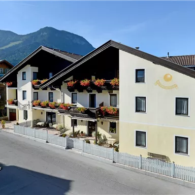 Ein gemütliches Gasthaus mit Blumenkästen und Balkonen. Im Hintergrund sind Berge und ein blauer Himmel zu sehen.