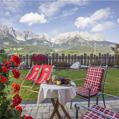 Eine gemütliche Terrasse mit Blick auf majestätische Berge. Im Vordergrund stehen blühende Blumen und ein Tisch mit frischen Früchten.