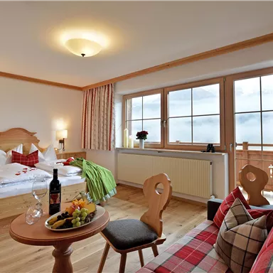 Ein gemütliches Hotelzimmer mit einem Doppelbett und einem Sitzbereich. Große Fenster bieten viel Tageslicht und den Blick auf die Berge.