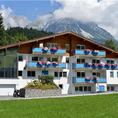 Ein modernes Gebäude mit Balkonen und blühenden Pflanzen. Im Hintergrund sind hohe Berge und ein blauer Himmel zu sehen.