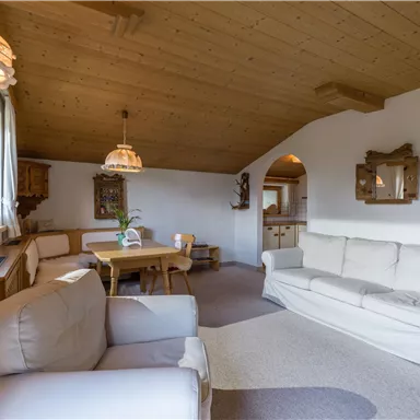 A cozy living room with a wooden ceiling and a large window. There is a bright couch, a dining table, and decorative elements on the walls.
