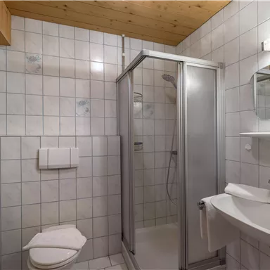 A bathroom with tiled walls, a shower, and a sink. The ceiling is made of wood, and there is a toilet in the room.