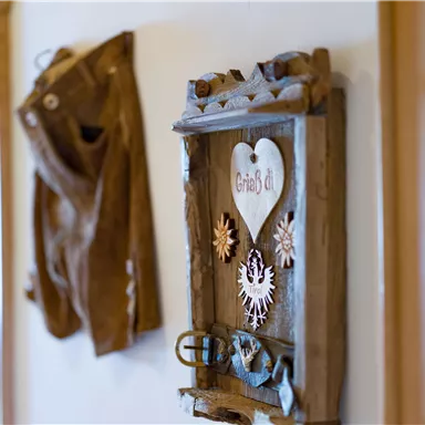 A rustic wall decor with a wooden frame and various ornamental elements. Next to the frame hangs a traditional leather trousers.