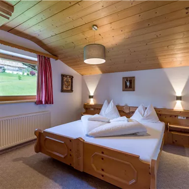 A cozy bedroom with wooden decor, two beds, and a small table. The window offers a view of greenery.