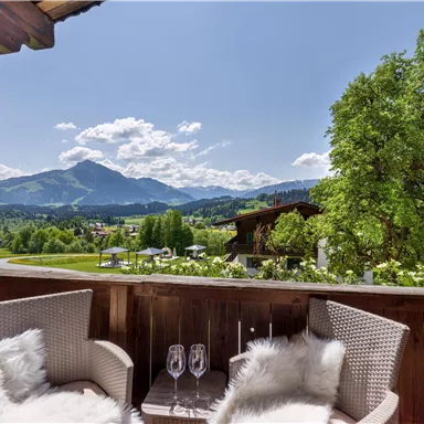A beautiful balcony with two comfortable chairs and a view of the surrounding mountains and the greenery of the landscape. The sky is clear and blue, perfect for relaxing moments outdoors.