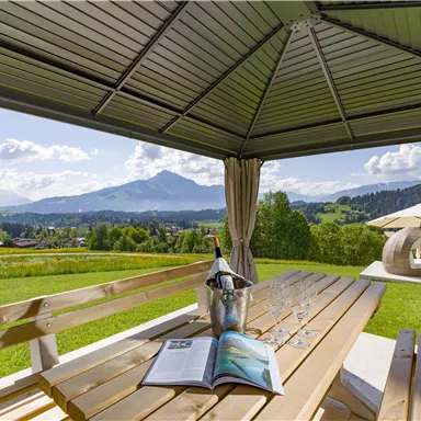 Ein gemütlicher Tisch unter einem Pavillon mit Blick auf die Berge. Auf dem Tisch stehen eine Flasche Sekt und Gläser, umgeben von einer schönen Landschaft.