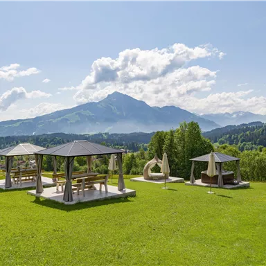 Eine grüne Wiese mit Blick auf die Berge und einem klaren Himmel. Im Vordergrund stehen Pavillons und entspannende Sitzmöglichkeiten.