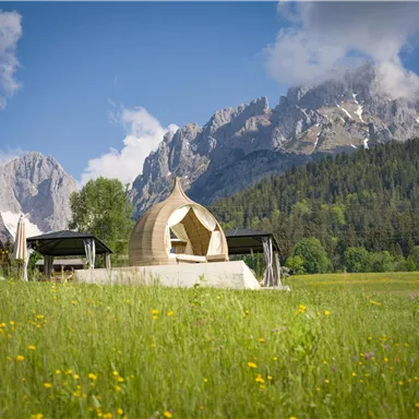 Eine moderne Struktur in einer grünen Wiese, umgeben von majestätischen Bergen. Der Himmel ist klar mit einigen Wolken.