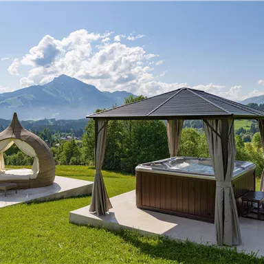 Ein entspannender Außenbereich mit einem Whirlpool und einem gemütlichen Sitzplatz. Im Hintergrund sind die Berge und ein klarer Himmel zu sehen.