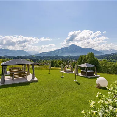 A green meadow with modern pavilions and an impressive mountain landscape in the background. The sky is clear and sunny, ideal for relaxation outdoors.