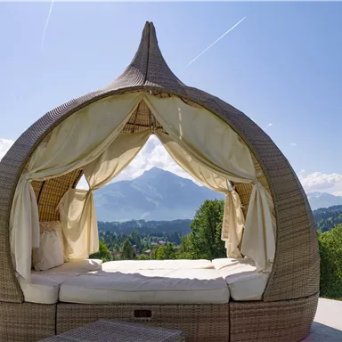 A cozy, stylish outdoor bed with a view of the mountains. The sky is clear and the surroundings are green and inviting.