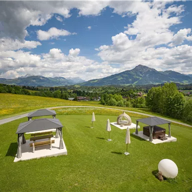 A picturesque landscape with green meadows and mountains in the background. In the foreground, there are cozy pavilions and seating areas.