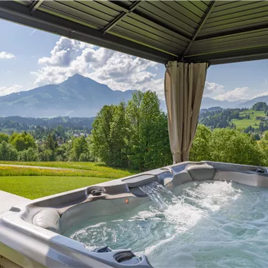 A relaxing hot tub under a canopy with a view of the mountains. The landscape is green, and the weather is sunny with some clouds.