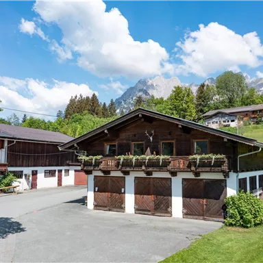 A traditional farmhouse surrounded by green meadows and mountains. The sky is blue with some white clouds.