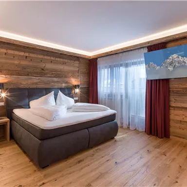 A modern bedroom with a large bed and wood paneling. A television displays an image of mountains, and there are windows with curtains.