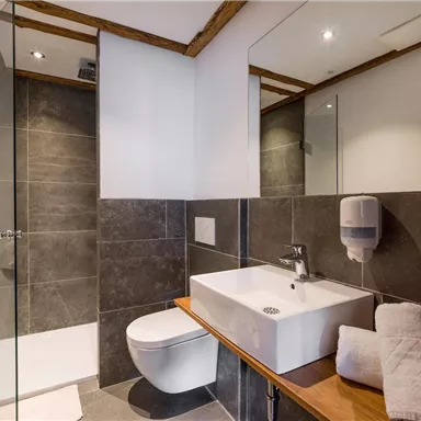 A modern bathroom with a clear shower and a simple washbasin. There are towels and a soap dispenser available, as well as elegant wood details.