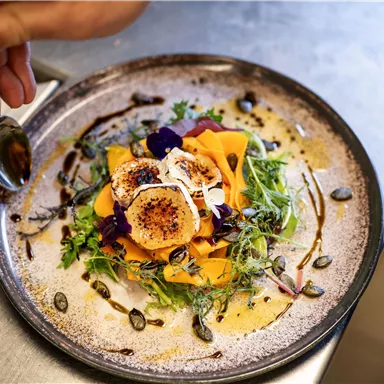 An artfully arranged salad on a dark plate. The salad contains colorful vegetable strips, herbs, and edible flowers.