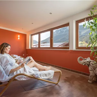 A woman relaxes in a deck chair and reads a book in a cozy room. Large windows offer a view of the mountains in the background.