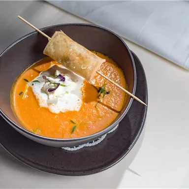 A delicious carrot soup with a piece of fried dough and a cream topping. Served in an elegant bowl on a dark plate.