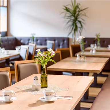 A cozy restaurant with wooden tables and plants. The tables are set for guests and decorated with flowers.