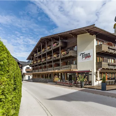 A charming hotel with wooden balconies and a beautiful facade. The street leads directly to the entrance, surrounded by green trees.