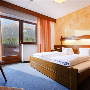 Ein gemütliches Schlafzimmer mit zwei Betten und einem Holzschreibtisch. Große Fenster mit Vorhängen bieten viel Tageslicht und einen Blick nach draußen.