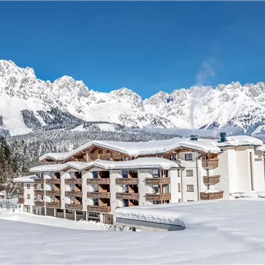 Ein malerisches Hotel inmitten einer schneebedeckten Landschaft mit Bergen im Hintergrund. Die klare, blaue Himmelsfarbe ergänzt die winterliche Szene.