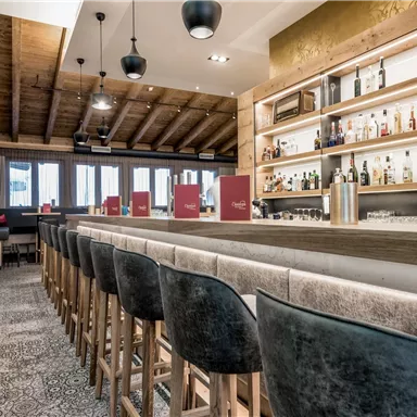 A modern bar with a long counter and stylish chairs. The walls are paneled with wood, and shelves display a selection of drinks.