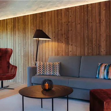 A modern living room with a gray sofa and a red armchair. Warm wooden walls and soft lighting create a cozy atmosphere.