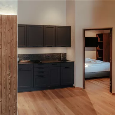 A modern kitchen with dark cabinets and wood paneling. In the background, a bedroom with a bed is visible.