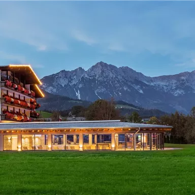A modern hotel with a large grassy area in the foreground and impressive mountains in the background. The facade of the hotel is illuminated, creating a welcoming atmosphere.